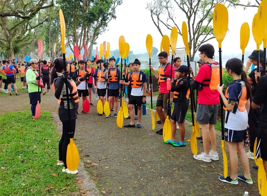 A group of young people wearing orange life jackets hold yellow kayak paddles outdoors near a waterway.